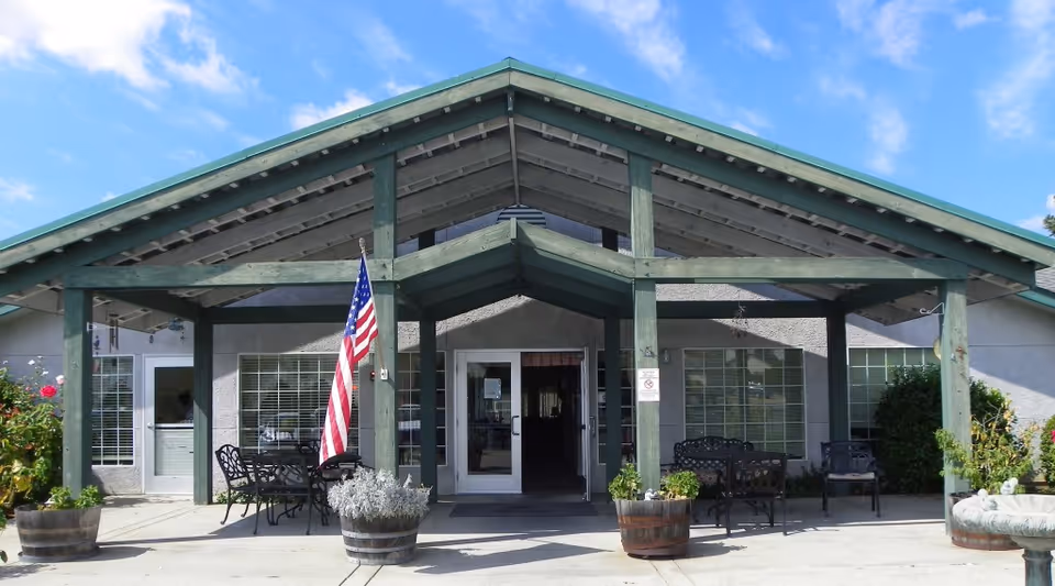 Front exterior view of a single-story building with a green wooden awning supported by beams. There are large windows on either side of the entrance door, outdoor metal chairs and tables, potted plants, an American flag, and a clear blue sky above.
