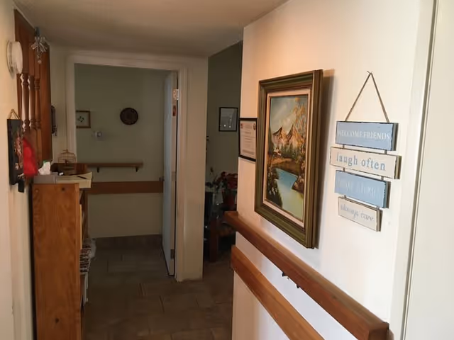 A hallway inside a senior living facility with beige tiled floor and white walls. On the right wall, there is a framed painting of a mountain landscape and a decorative hanging sign with the words 'WELCOME FRIENDS, laugh often, SHARE STORIES, always care.' Wooden handrails run along both sides of the hallway. At the end of the hallway, there is an open door leading to another room with a bench and wall decorations.