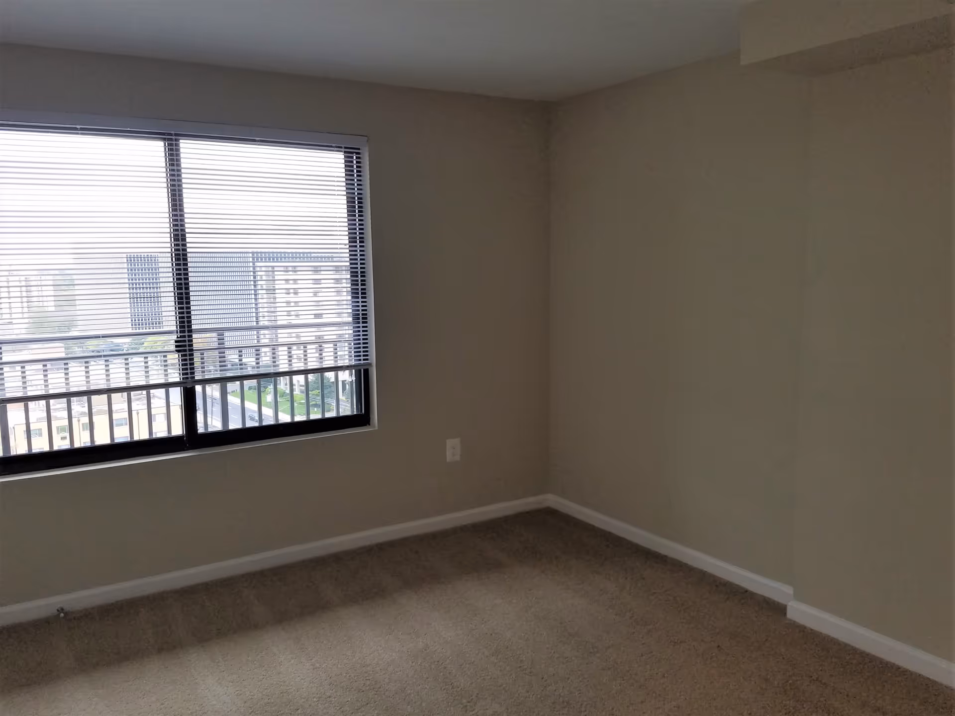 Empty room with beige carpet and walls, a large window with horizontal blinds partially open showing a view of buildings outside.