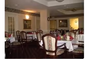 Dining room with multiple tables covered in white tablecloths and set with folded napkins and glassware. The room has upholstered chairs with wooden frames, framed artwork on the walls, and soft lighting fixtures.