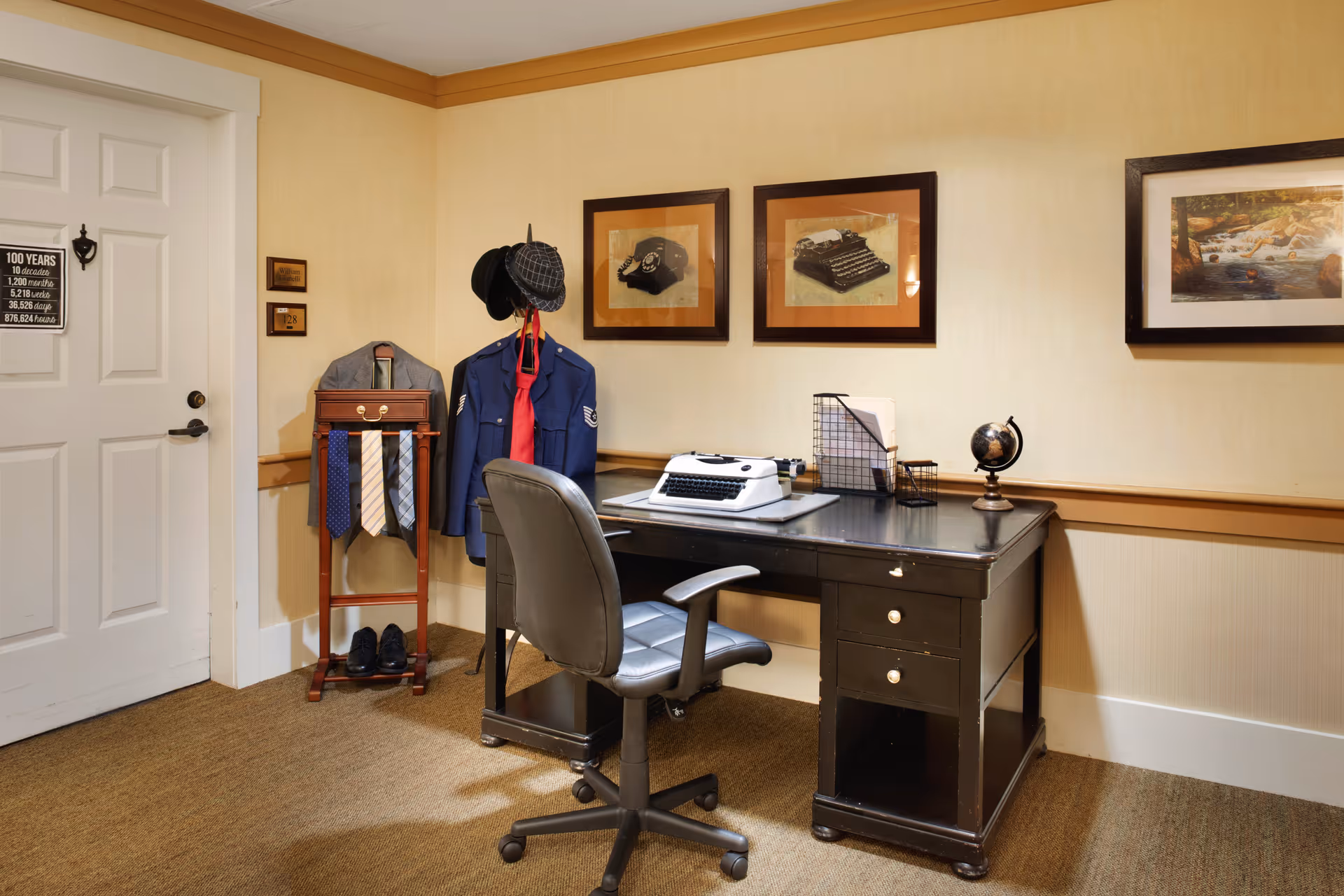 A cozy office corner with a black desk and a black swivel chair. On the desk, there is a white typewriter, a small globe, and a wire organizer with papers. Behind the desk, two framed pictures of a vintage telephone and a typewriter hang on the beige wall. To the left, a coat rack holds a blue uniform jacket with a red tie, a gray jacket, several neckties, and a pair of black shoes on the floor. A white door with a sign listing time increments for 100 years is visible on the left side of the image.