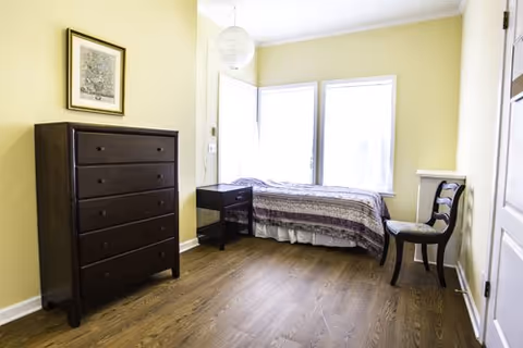 A small, simple bedroom with light yellow walls and wood flooring. The room contains a single bed with a patterned bedspread positioned near two windows with white curtains. There is a dark wooden dresser with five drawers on the left side, a small dark wooden nightstand next to the bed, and a wooden chair with a cushioned seat near the door on the right.