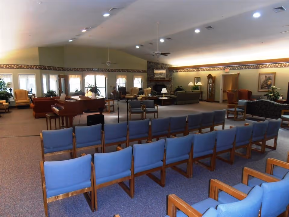 A spacious common area in a retirement community with rows of blue cushioned chairs arranged facing a piano and microphone stand. The room features multiple seating areas with armchairs and sofas, a stone fireplace, ceiling fans, and large windows letting in natural light.