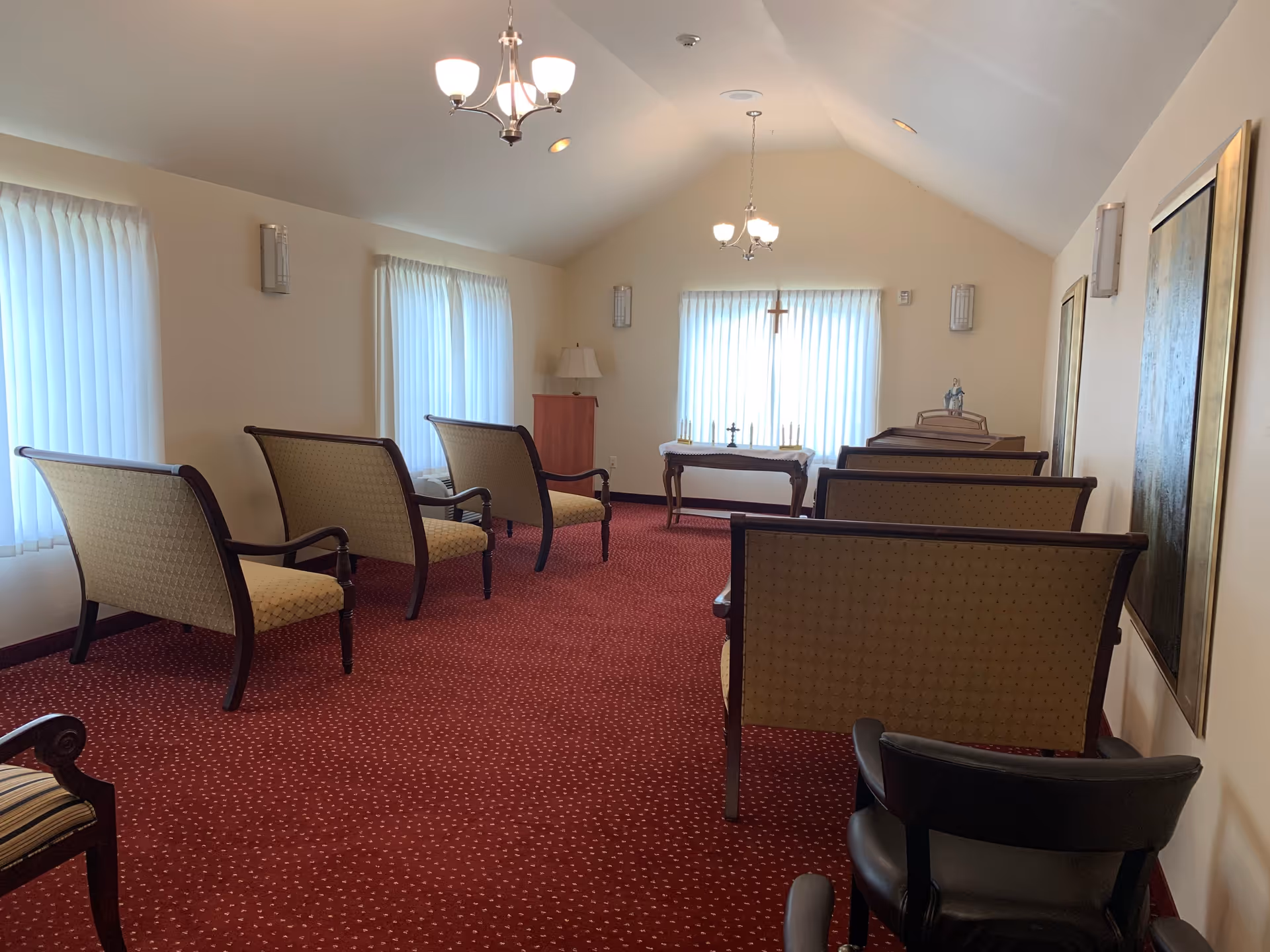 A small chapel or meditation room with several upholstered chairs arranged in rows facing a table with religious items, including a cross, candles, and a statue. The room has a red carpet, cream-colored walls, three windows with sheer white curtains, and two ceiling light fixtures.