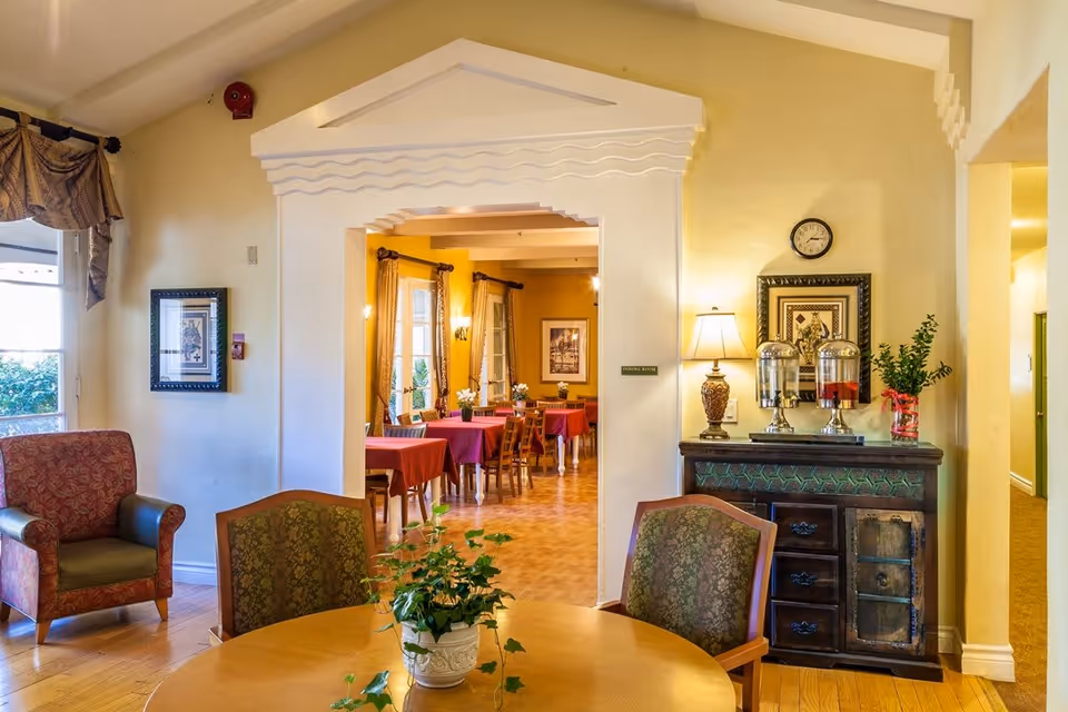 Interior view of a senior living facility showing a cozy seating area with a round wooden table and two upholstered chairs. A potted plant is on the table. In the background, there is an arched doorway leading to a dining room with tables covered in red tablecloths and wooden chairs. The walls are painted yellow, and there are framed pictures and a clock on the walls. A sideboard with a lamp, two beverage dispensers, and a vase with flowers is also visible.