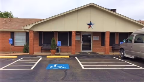 Front exterior view of Concho Health and Rehabilitation Center showing a single-story brick building with a beige gabled roof. There are two benches and potted plants near the entrance, a large star decoration above the door, and several handicap parking spaces in front.