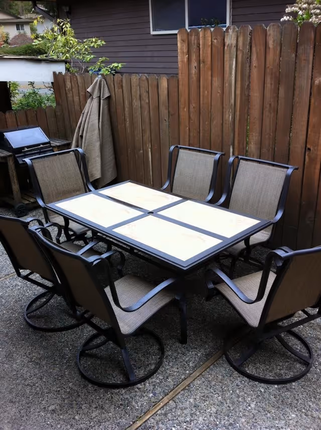 Outdoor patio area with a rectangular table featuring a tiled surface and six metal-framed chairs with mesh seats and backs. The area is enclosed by a wooden fence, and there is a barbecue grill and a hanging cloth or towel in the corner.