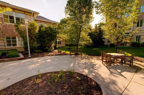 Sunny landscaped courtyard with patio seating, trees, and surrounding brick buildings.