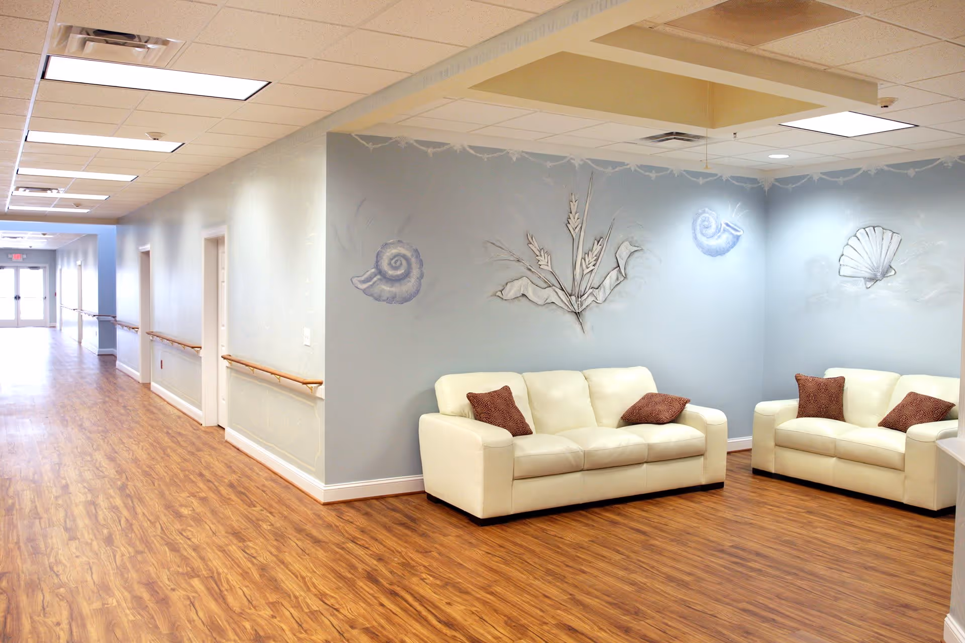 A bright hallway in a senior living facility with wooden flooring and light blue walls. Two white leather sofas with brown cushions are placed in a corner seating area. The walls are decorated with artistic designs of seashells and plants. Handrails run along the hallway walls, and fluorescent ceiling lights illuminate the space.