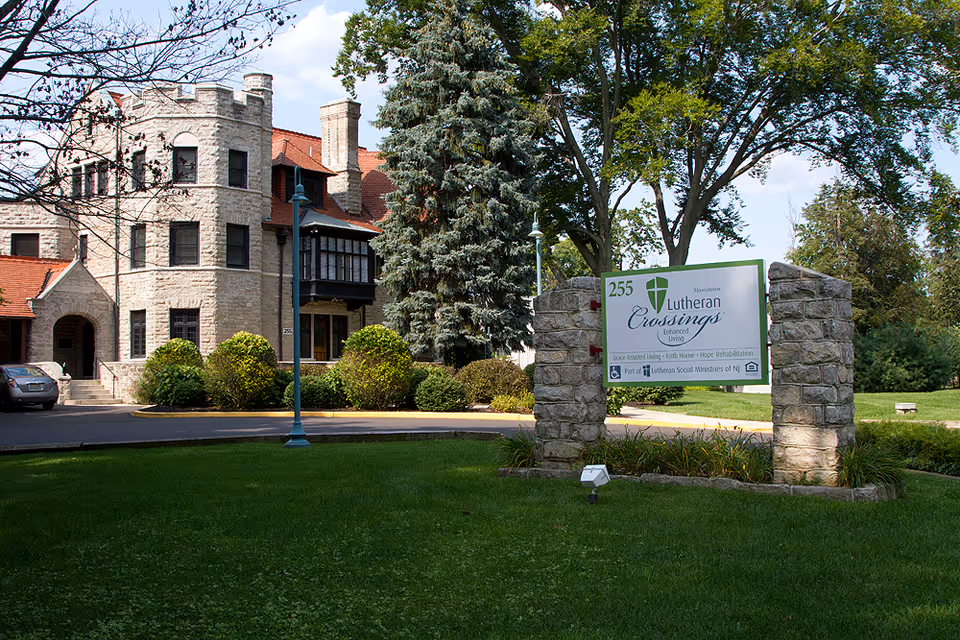 Exterior view of a stone building with a red roof surrounded by trees and bushes. In the foreground, there is a stone signpost with a green and white sign that reads '255 Lutheran Crossings Enhanced Living' along with additional text about assisted living and rehabilitation services.