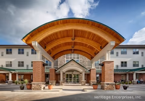 Front exterior view of Evergreen Senior Living - Decatur featuring a large covered entrance with a curved wooden ceiling supported by brick and stone pillars, potted plants along the driveway, and a multi-story building with numerous windows under a partly cloudy sky.