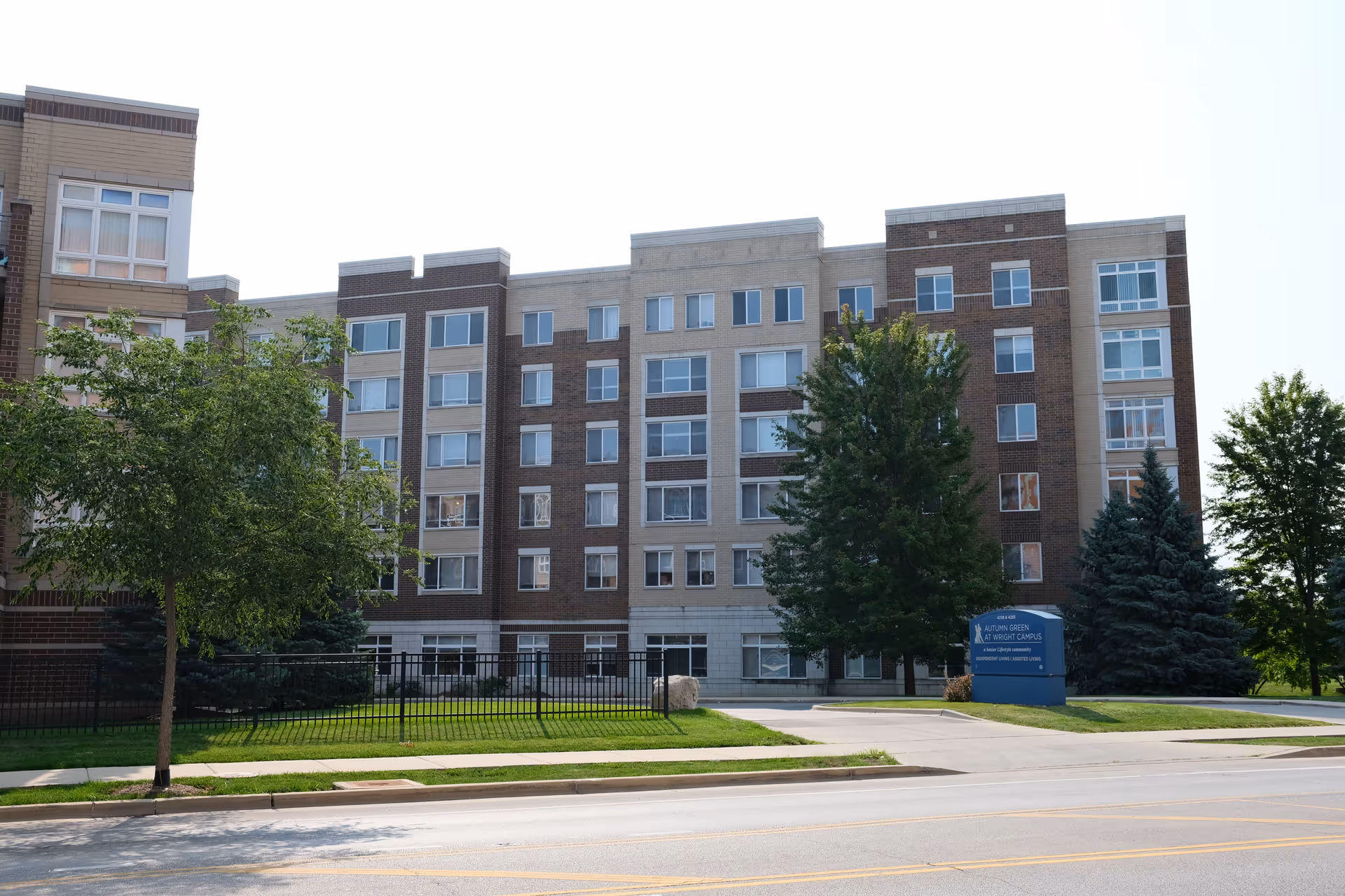 Exterior view of a multi-story senior living facility building with a mix of beige and brown brick facade, several windows, and trees in front. A blue sign near the entrance reads 'Autumn Green at Wright Campus' indicating it is a senior living community.