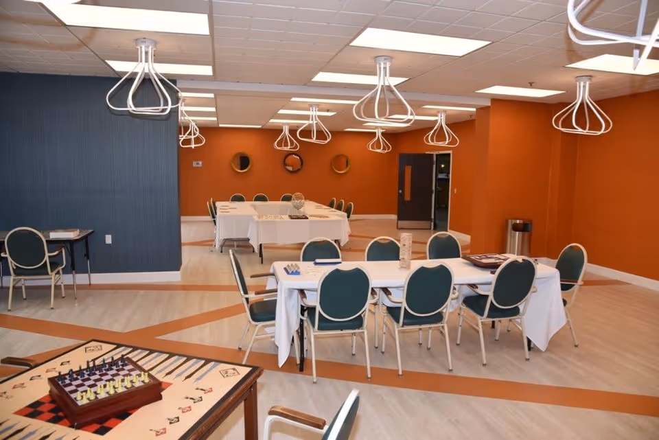 A spacious room with orange and blue walls, featuring several tables covered with white tablecloths and surrounded by green cushioned chairs. The tables have board games and puzzles on them. Modern white ceiling lights hang above, and there are three round mirrors on the far orange wall. The floor has a light wood pattern with orange accents.