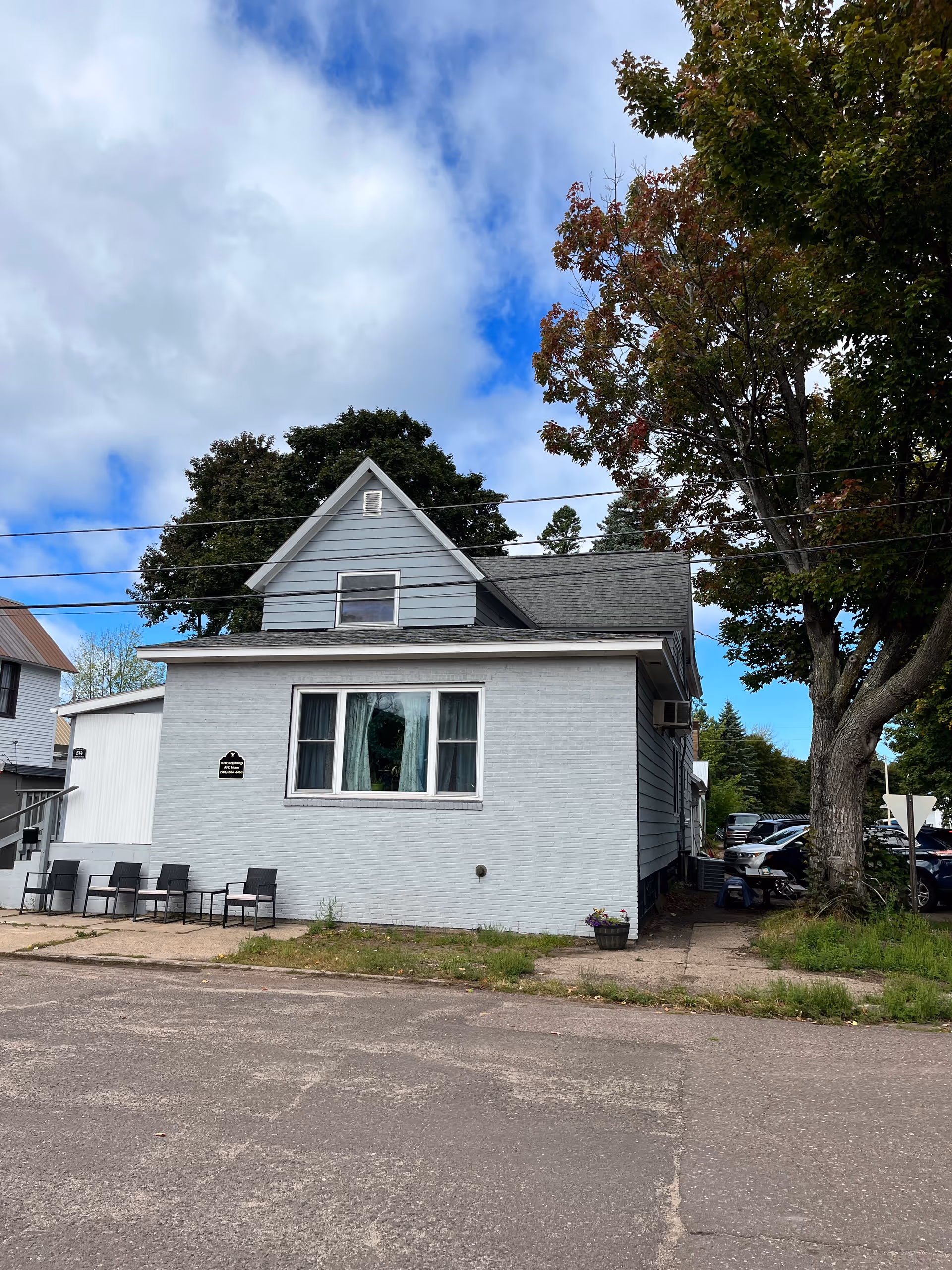 Exterior view of a two-story light gray building with a small patio area featuring several chairs and a table. There is a large tree to the right side and several parked cars visible behind the building. The sky is partly cloudy with patches of blue.