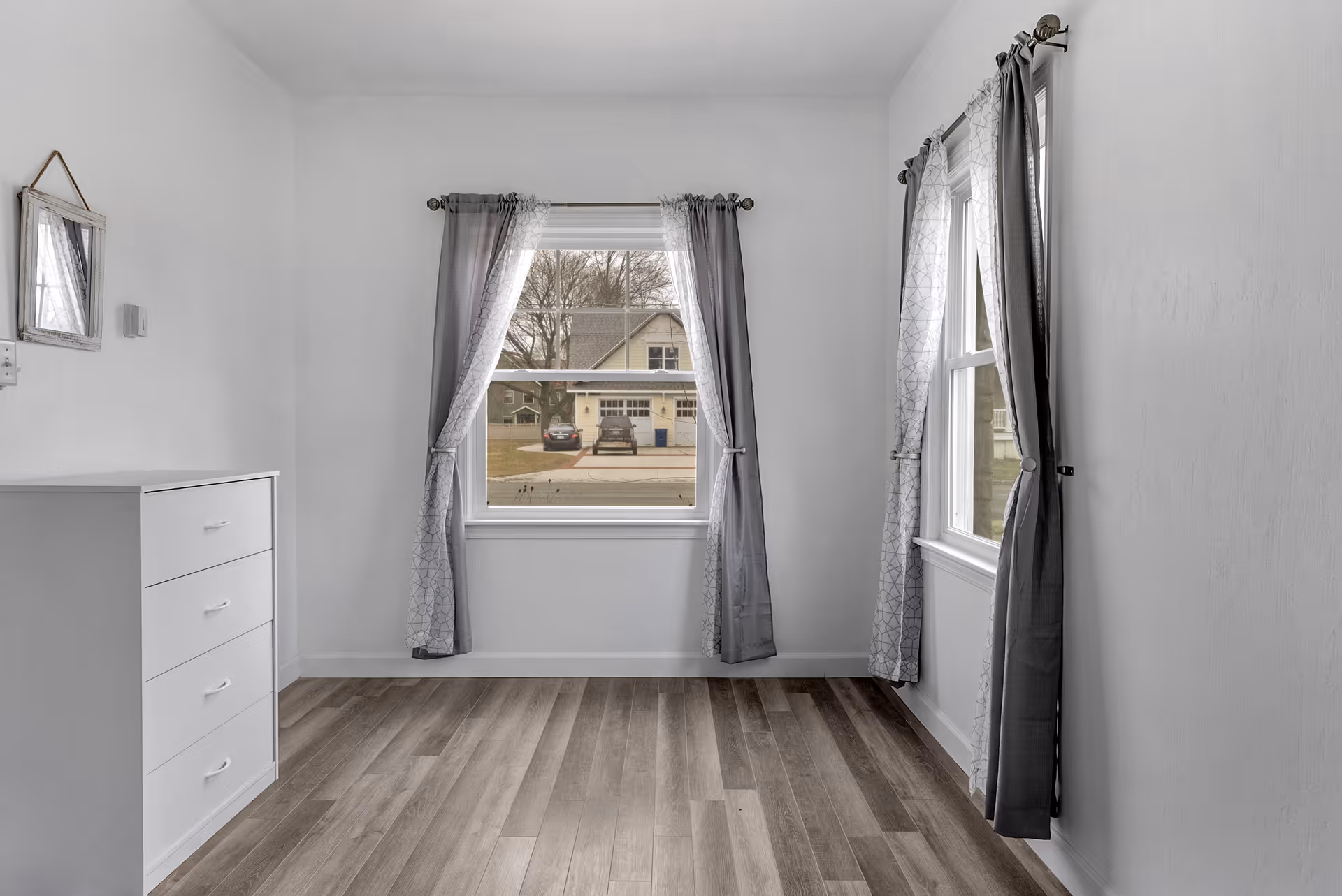 Empty room with two windows covered by gray curtains, a white chest of drawers on the left, a small mirror hanging on the wall, and wood-patterned flooring.