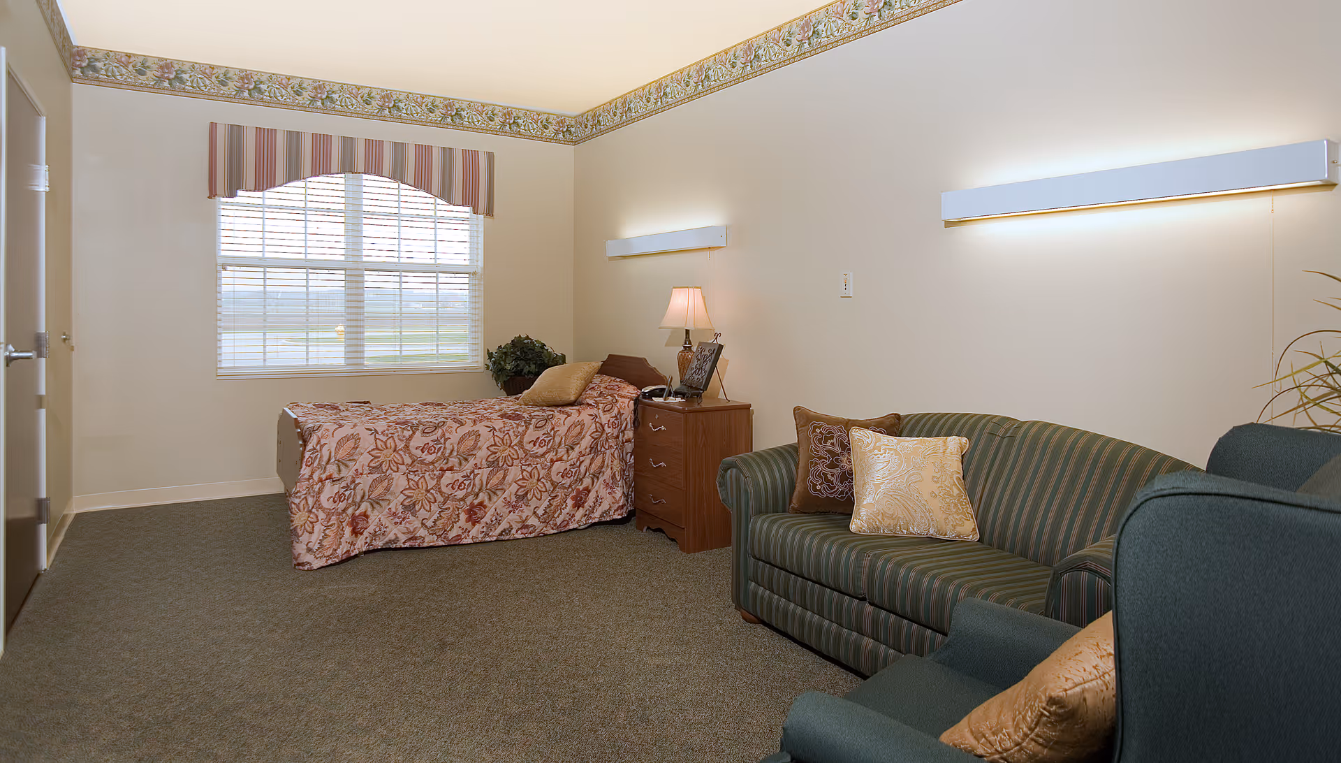 A senior living bedroom with a single bed, nightstand and lamp, plus a seating area with a striped sofa and armchair.