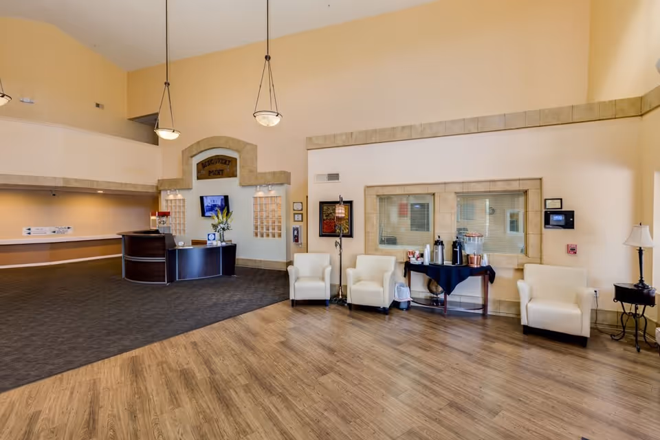 Spacious reception area of Discovery Point Retirement Community with a curved dark wood front desk, three white armchairs, a small table with coffee and refreshments, and warm beige walls with stone trim accents. The floor is a combination of wood and carpet, and two pendant lights hang from the high ceiling.