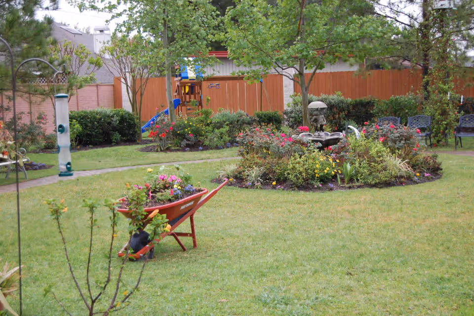A well-maintained garden area with a circular flower bed featuring various colorful flowers and a small fountain in the center. Surrounding the flower bed are green grass and several trees. A red wheelbarrow filled with flowers is positioned on the lawn. In the background, there is a wooden fence, a children's play structure with a slide, and several black metal chairs arranged along the fence.