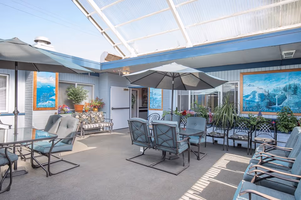 Outdoor patio area with multiple seating arrangements including cushioned chairs, a glass table with an umbrella, and a bench with floral cushions. The space is decorated with potted plants and two large landscape paintings on the walls. The area is covered with a translucent roof allowing natural light to enter.