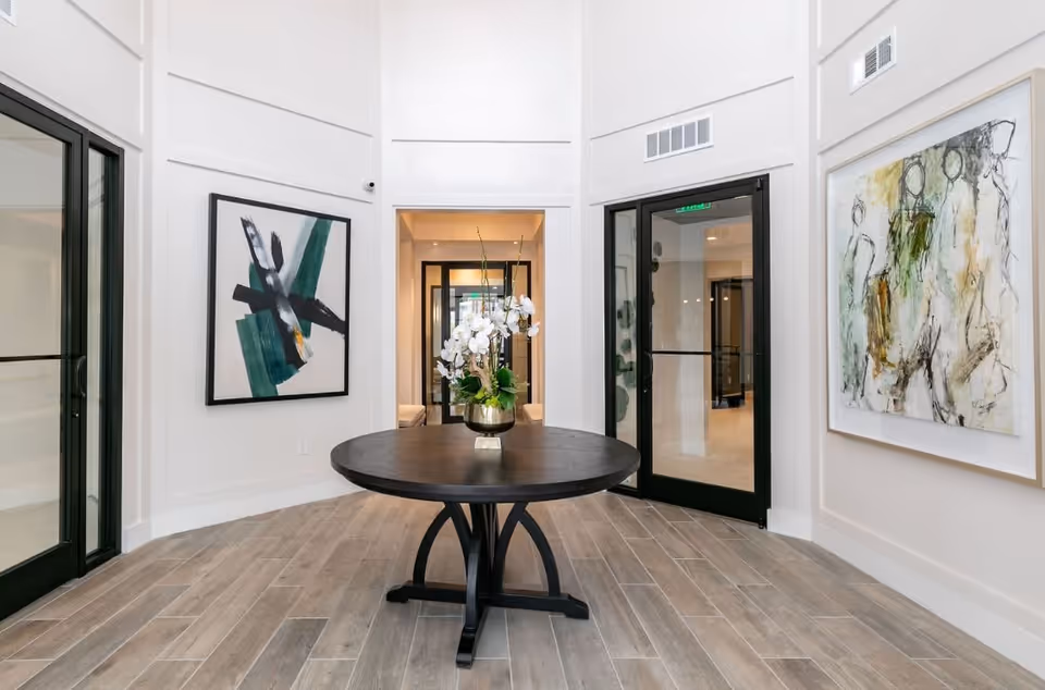 A modern interior space with a round black table in the center holding a vase with white flowers. The room has light-colored wooden flooring, white walls with decorative molding, and two abstract paintings on the walls. There are black-framed glass doors on either side and a hallway visible in the background.
