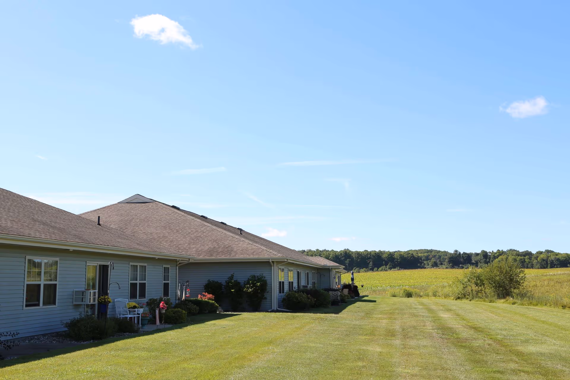 Single-story building with light blue siding and a brown shingled roof, surrounded by a well-maintained grassy lawn and some bushes, under a clear blue sky with a few clouds. The building is adjacent to open fields and trees in the distance.