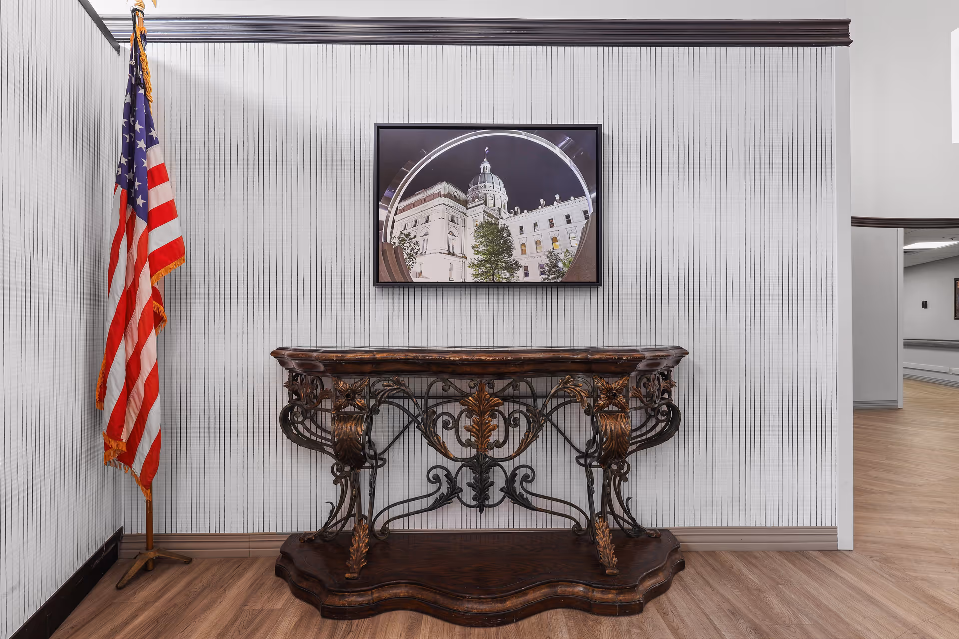 Ornate wooden console table against a patterned wall with a framed photo above and an American flag standing to the left in a corridor.