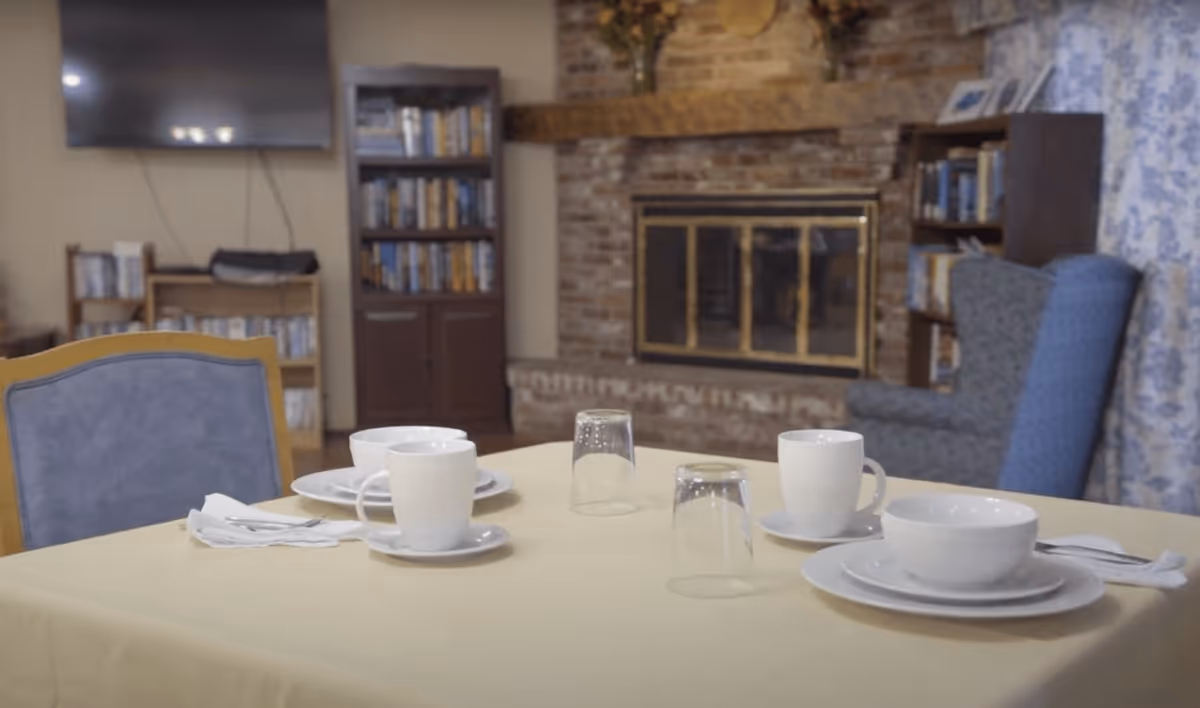 A dining table set with white cups, bowls, plates, and glasses in a cozy room. In the background, there is a brick fireplace with a wooden mantle, bookshelves filled with books, a blue upholstered chair, and a wall-mounted television.