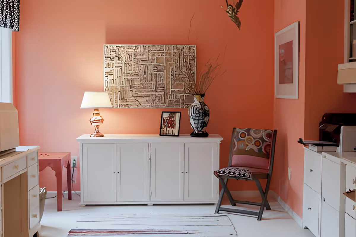 A cozy interior corner with coral-colored walls featuring a white cabinet topped with a decorative lamp, a framed photo, and a large artistic vase with dried branches. To the right is a wooden chair with a colorful cushion, and above the cabinet hangs a textured abstract artwork. The floor is light-colored with a subtle rug, and part of a kitchen counter is visible on the right.
