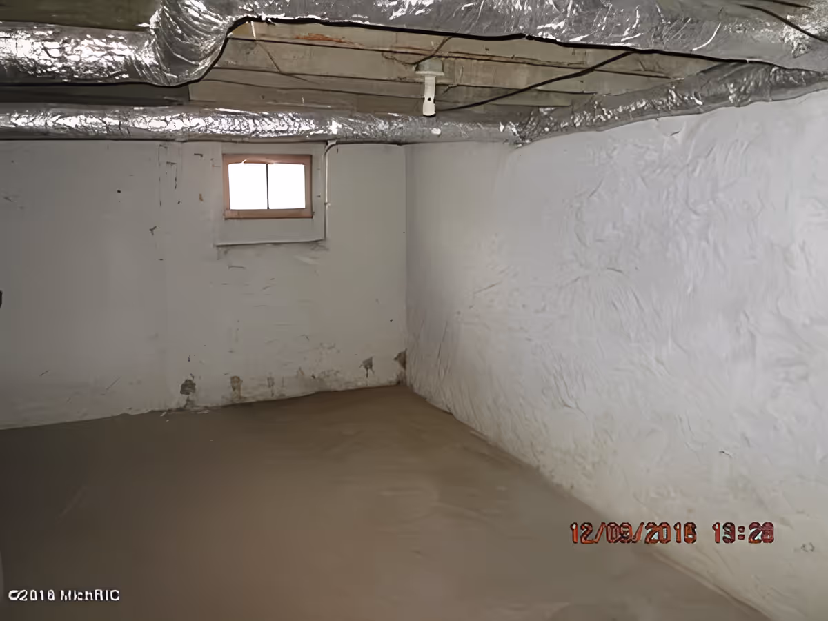 Empty basement room with white textured walls, a small window near the ceiling, exposed ductwork wrapped in insulation on the ceiling, and a concrete floor.