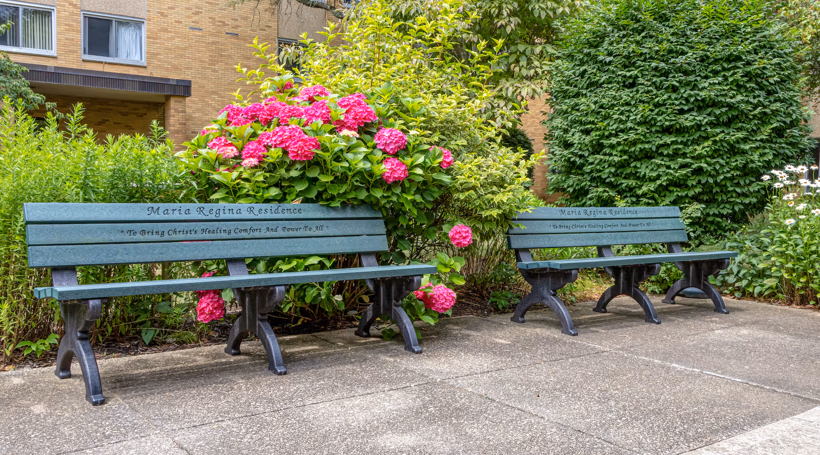 Two green benches with black legs placed on a concrete pathway in front of a garden with vibrant pink flowers and green foliage. The benches have the inscription 'Maria Regina Residence' and the phrase 'To Bring Christ's Healing Comfort And Power To All.' A brick building is partially visible in the background.