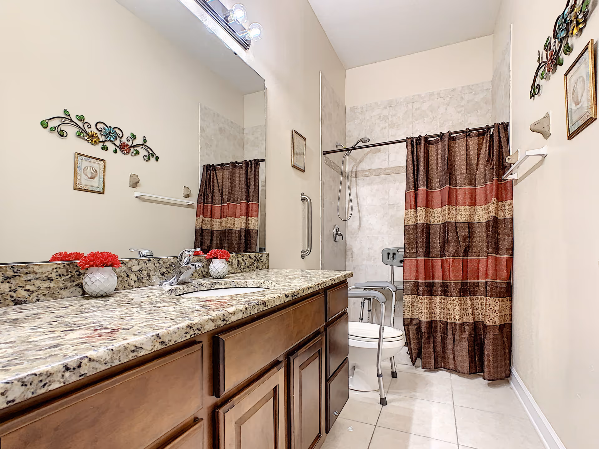 A bathroom with a granite countertop and wooden cabinets. There is a large mirror above the sink, decorated with a small vase of red flowers. The bathroom features a toilet with a walker in front of it, a shower with a brown and red striped shower curtain, and a grab bar on the wall next to the shower. The walls are light-colored and decorated with a floral metal wall art and framed pictures.
