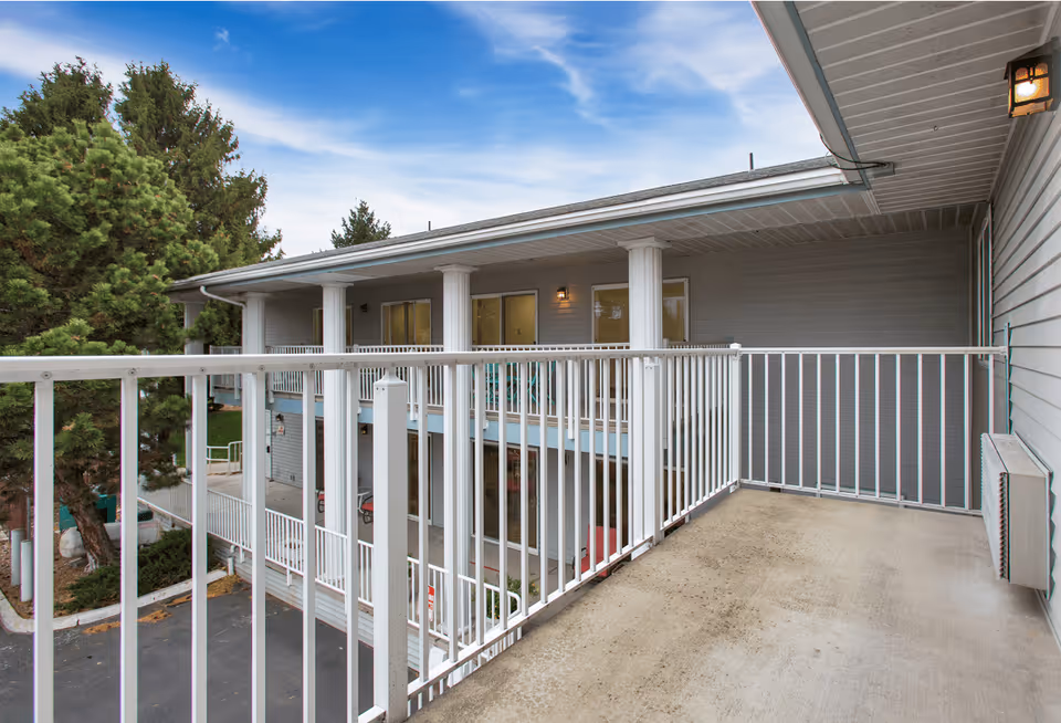 View of an outdoor balcony area with white metal railings attached to a senior living facility building. The balcony overlooks a parking area and is adjacent to a gray building with multiple windows and white columns. Trees and a partly cloudy sky are visible in the background.