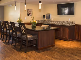Interior view of a dining area with a long counter and several high chairs. The counter has a dark granite top and wooden base. There are pendant lights hanging from the ceiling, a wall-mounted TV, and a paper towel dispenser on the wall. The floor is wooden, and there are decorative plants on the counter.