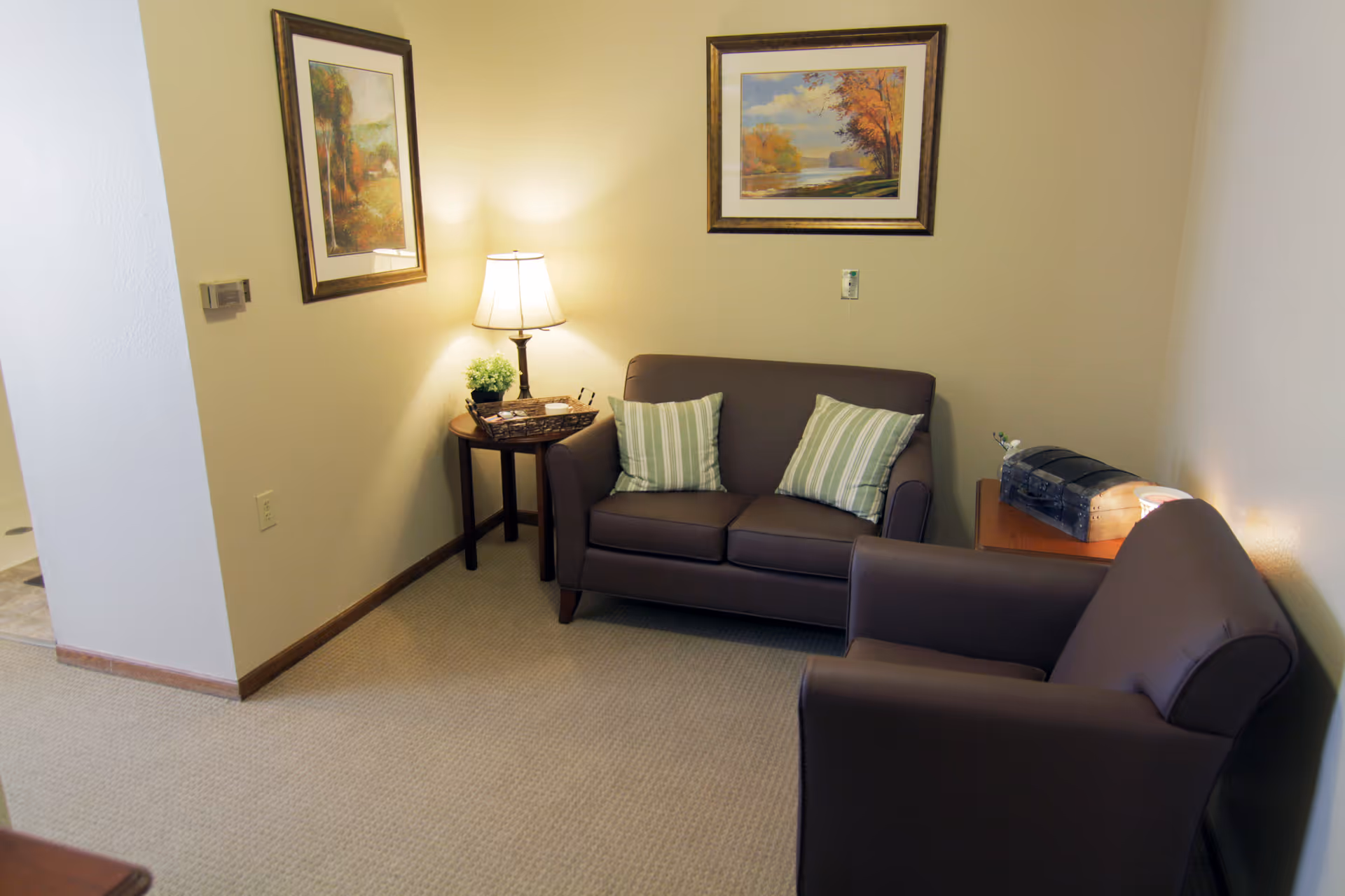 Cozy sitting area with a brown loveseat and armchair, side tables with a lamp and decorative items, and framed artwork on beige walls.