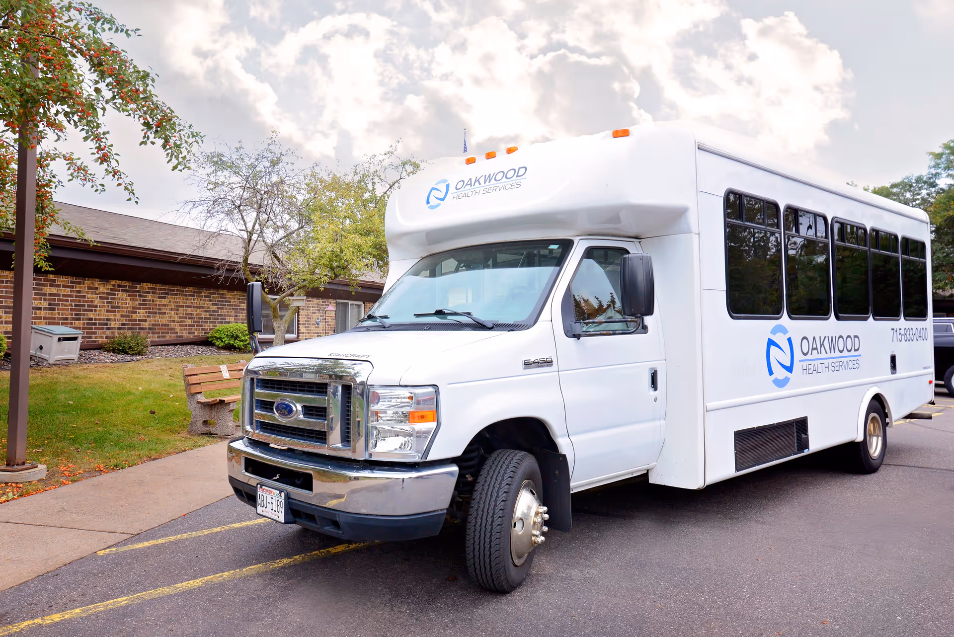 A white Oakwood Health Services shuttle bus parked outside a brick senior living building with a bench and trees.