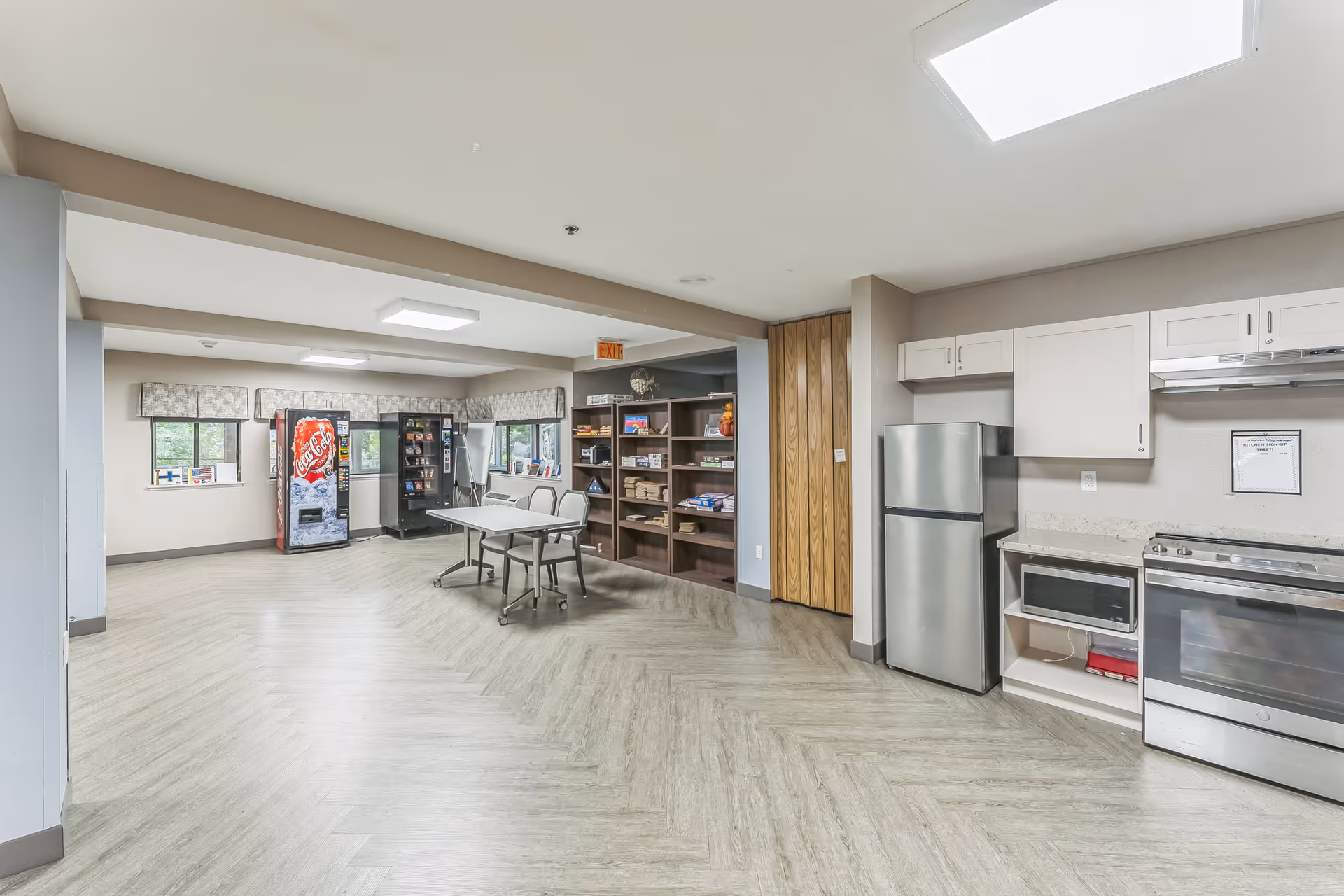 Interior view of a common area in a senior living facility featuring a kitchenette with a refrigerator, microwave, and stove on the right. In the background, there are vending machines, a table with chairs, and shelves stocked with various items. The room has light-colored walls, wood-patterned flooring, and ceiling lights.