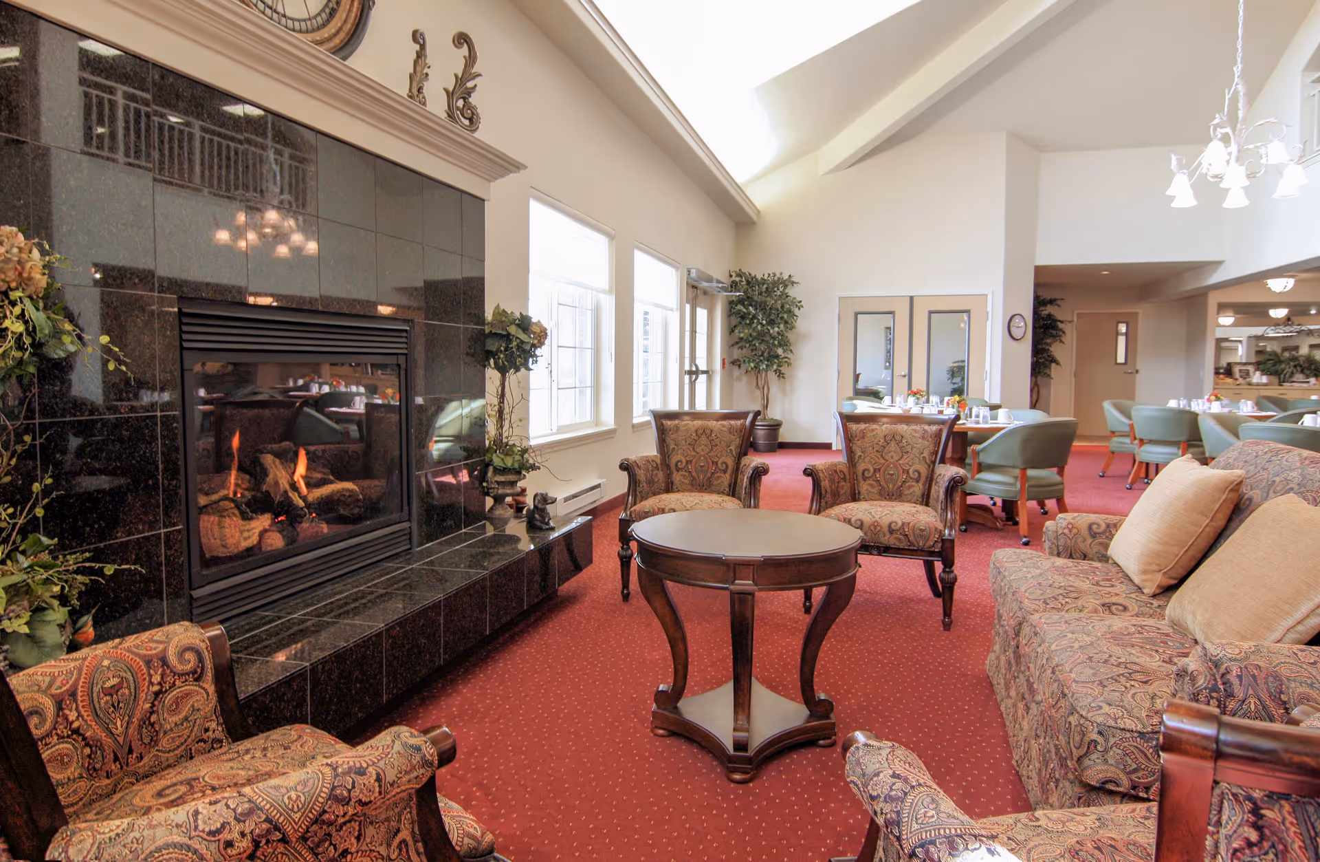 A cozy living room area with a lit fireplace surrounded by black tiles. The room features patterned upholstered armchairs and a sofa with beige cushions, a round wooden coffee table, and a red carpet. Large windows allow natural light to fill the space, and there are dining tables and chairs visible in the background.