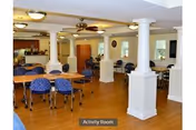 Spacious activity room with wooden flooring, multiple tables and blue cushioned chairs, white columns, ceiling fans, and windows allowing natural light.