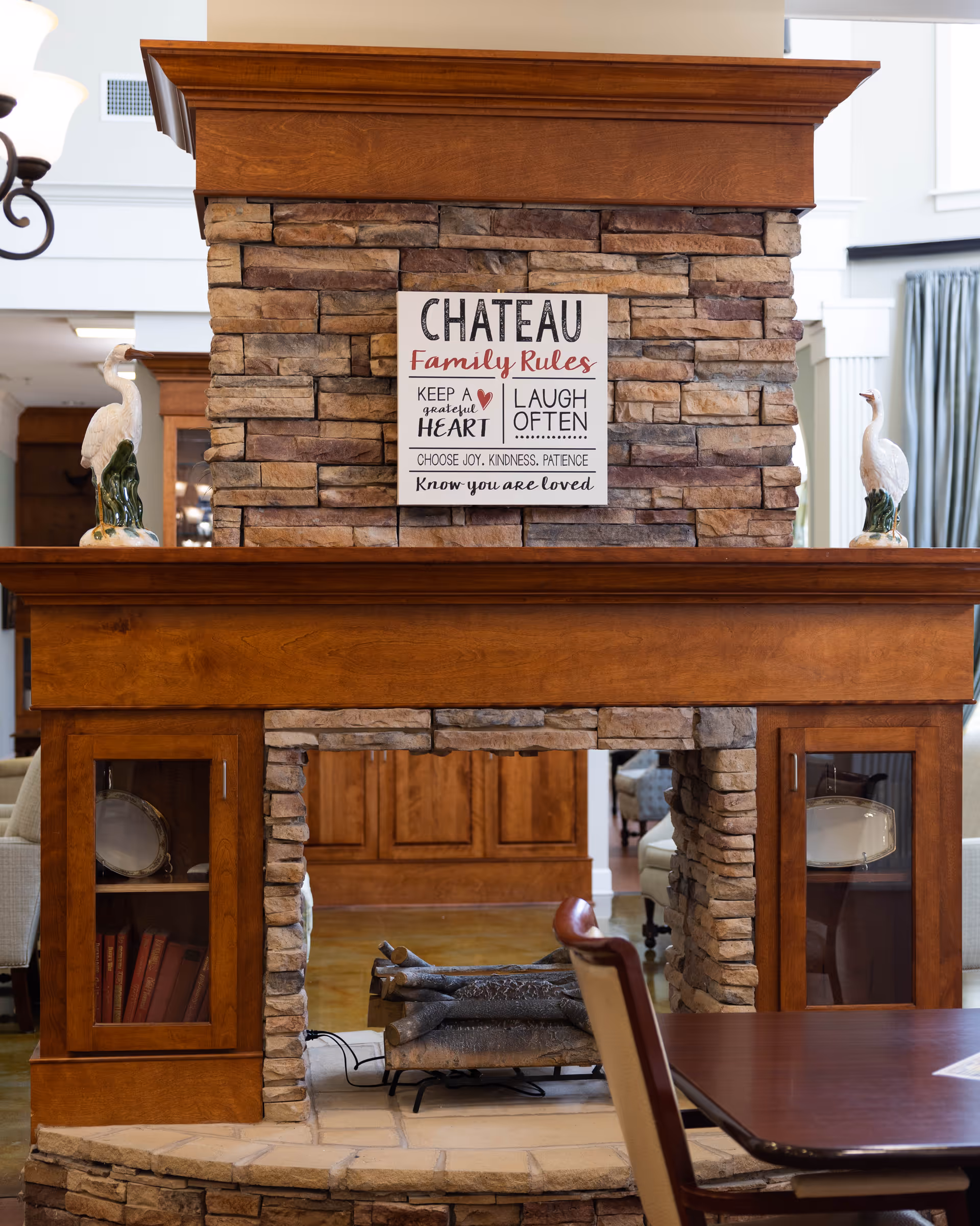 A stone and wood fireplace with a sign that reads 'CHATEAU Family Rules: Keep a grateful heart, Laugh often, Choose joy, kindness, patience, Know you are loved.' Two white bird figurines are placed on either side of the fireplace mantel. In the foreground, there is a wooden table and chair, and the background shows part of a cozy interior with wooden cabinets and curtains.