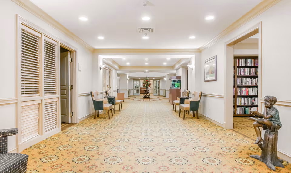 A wide, well-lit hallway in a senior living community with patterned carpet, several chairs along the walls, a decorative table with flowers at the far end, an aquarium, and a bronze statue of a boy reading a book near a doorway leading to a room with bookshelves.