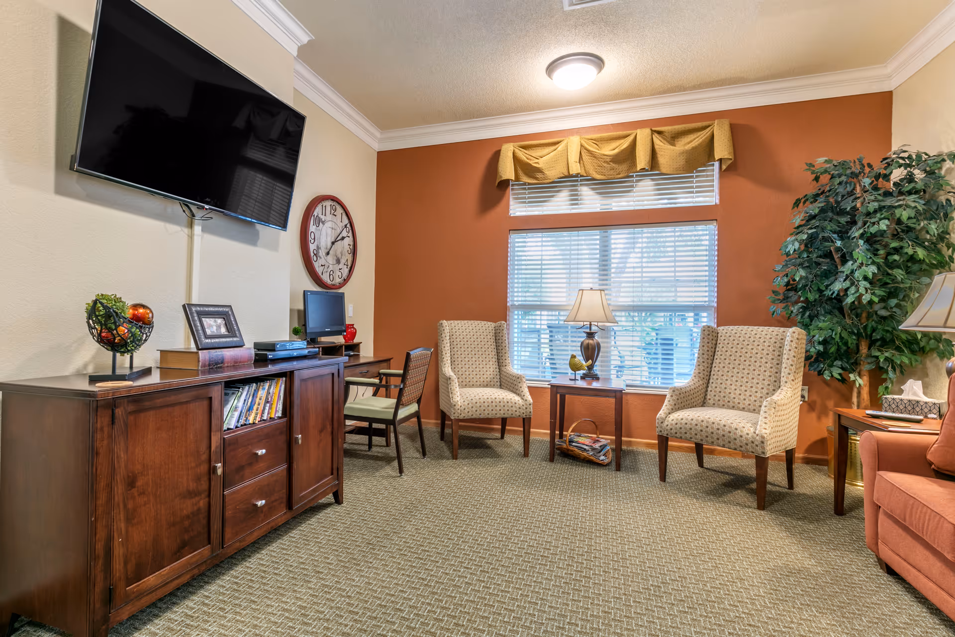 A cozy living room area with two patterned armchairs and a small wooden table with a lamp and decorative bird figurine in front of a window with blinds and a yellow valance. To the left, there is a wooden cabinet with books, a decorative bowl with fruit, a framed photo, and a flat-screen TV mounted on the wall above. A large round clock hangs on the wall next to a small desk with a chair and computer monitor. A tall artificial plant is placed in the corner next to a side table with a lamp and tissue box.