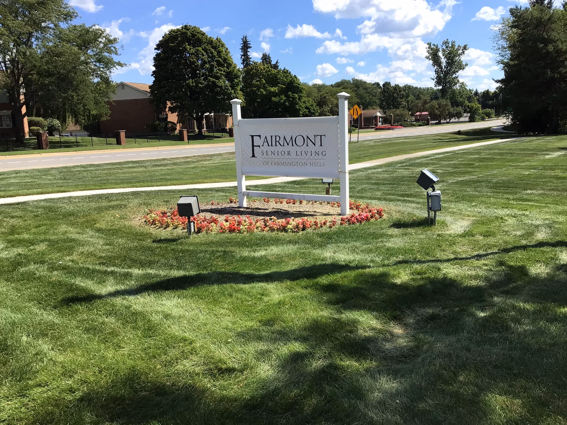 A white sign with black text reading 'Fairmont Senior Living of Farmington Hills' is displayed on a grassy area with flower beds around its base. The background shows a road, trees, and a partly cloudy sky.