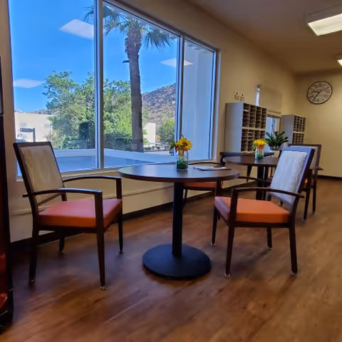 A bright room with large windows showing an outdoor view with palm trees and hills. Inside, there are round tables with two chairs each, decorated with small flower vases. The room has wooden flooring, a clock on the wall, and mail cubbies in the background.