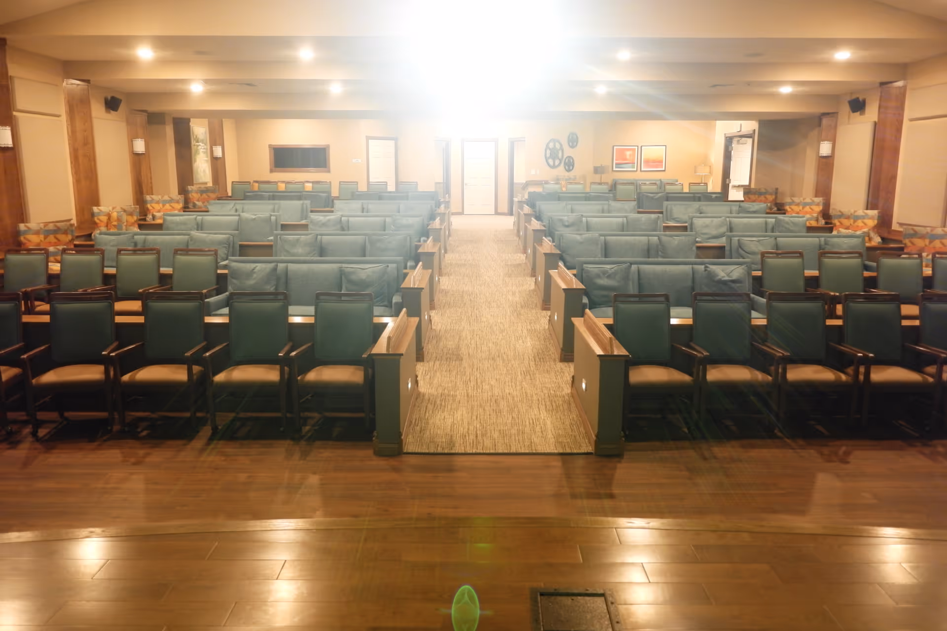 Interior view of a large room with multiple rows of green cushioned chairs arranged in a theater or auditorium style, facing a stage area with wooden flooring. The room has beige walls, wooden pillars, and soft lighting from ceiling fixtures.