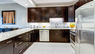Modern open kitchen with dark wood cabinets, marble countertops, stainless steel refrigerator and dishwasher, and a breakfast bar.