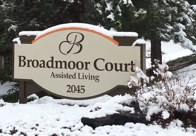 A large outdoor sign covered with a light layer of snow that reads 'Broadmoor Court Assisted Living 2045' surrounded by snow-covered ground, bushes, and trees.