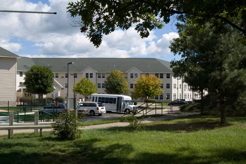 Exterior view of a senior living facility named Village at the Oaks with a parking lot, several cars, a white shuttle bus, trees, and a grassy area under a partly cloudy sky.