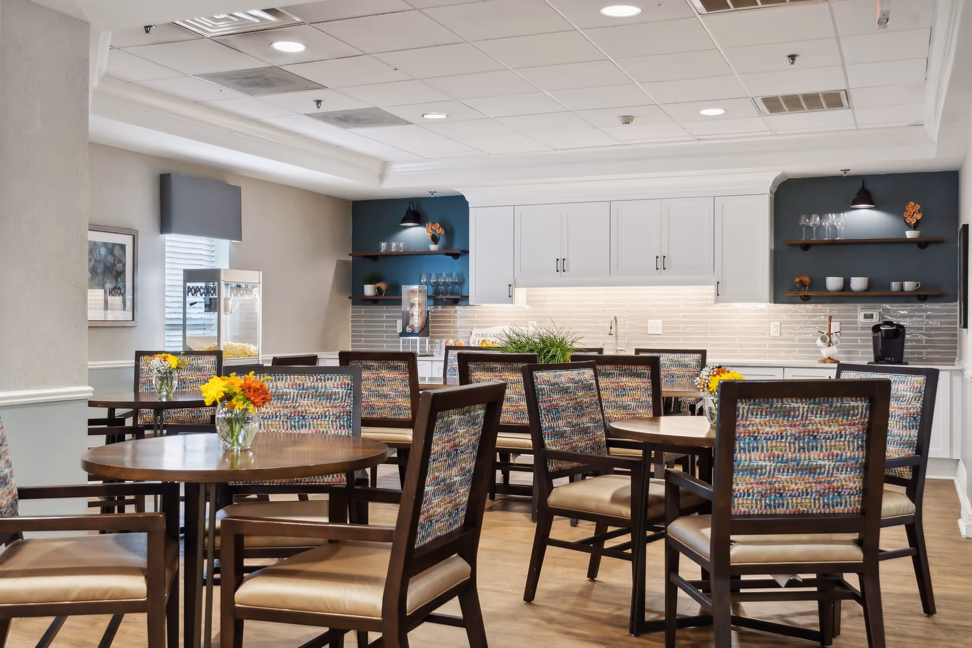 Communal dining room with round wooden tables, patterned upholstered chairs, floral centerpieces, and a kitchen counter in the background.