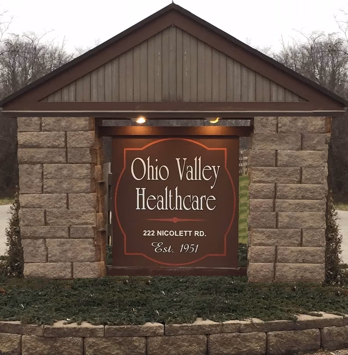 Outdoor stone sign structure with a peaked roof displaying the name Ohio Valley Healthcare, the address 222 Nicolett Rd., and the establishment year 1951, surrounded by greenery and trees in the background.