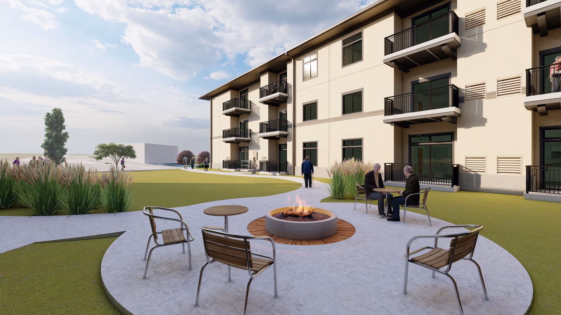 Outdoor seating area with a circular fire pit surrounded by metal and wooden chairs on a concrete patio. Two elderly people are sitting and conversing near the fire pit. The area is adjacent to a multi-story building with balconies and large windows. There are landscaped grassy areas and plants around the patio, with a partly cloudy sky overhead.